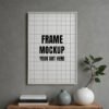 Frame mockup PSD showcasing a white-framed artwork with a grid design displayed on a gray wall, alongside a table with decorative vases and stacked books.