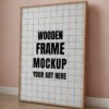 A wooden frame mockup with a blank interior leaning against a soft pink wall, on a wooden floor.