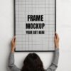 Frame mockup PSD featuring a person holding a large black-framed empty in front of a white wall.