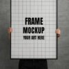 Frame mockup PSD featuring a person holding a large black-framed empty canvas against a gray brick wall.